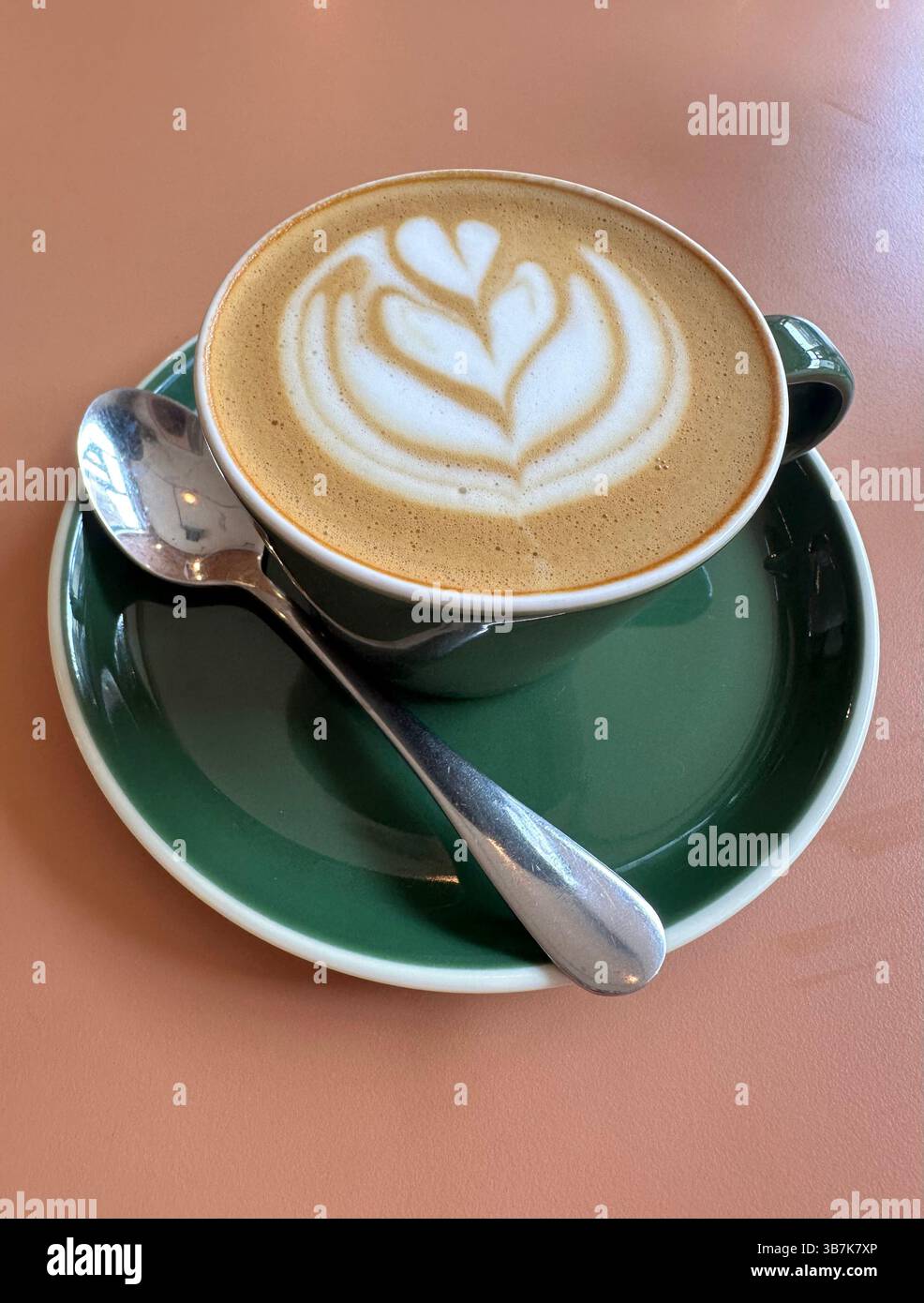 Flat white coffee in green cup and saucer with spoon on peach table, side view. - Smartphone Captured Stock Image