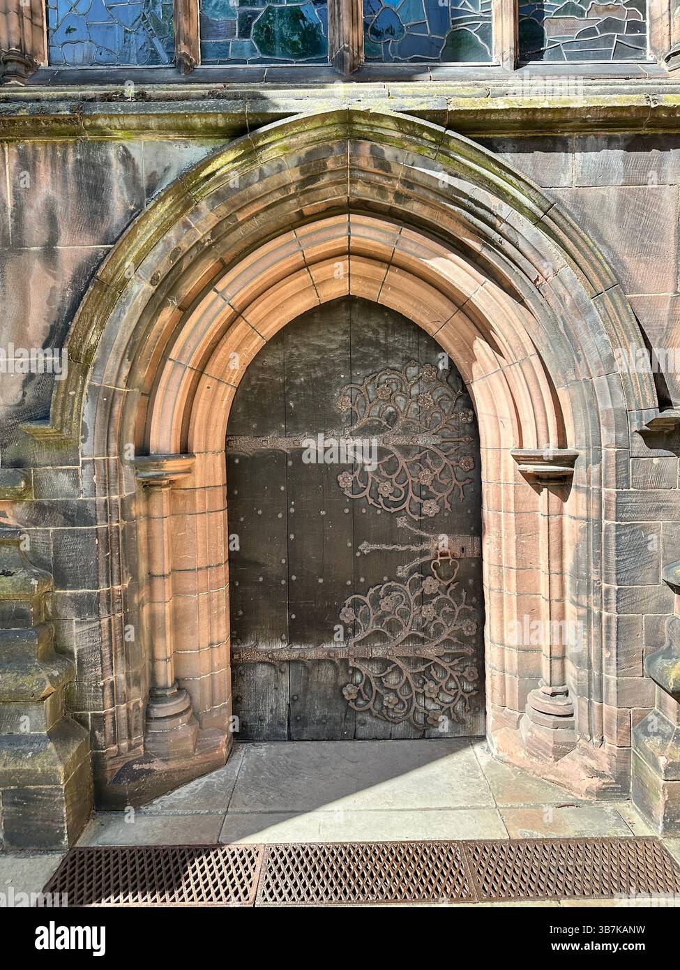 Ornate small wooden door with iron fixings at Chester Cathedral, framed by red brick arch and stained glass above. Taken on a sunny day with shadows. - Smartphone Captured Stock Image