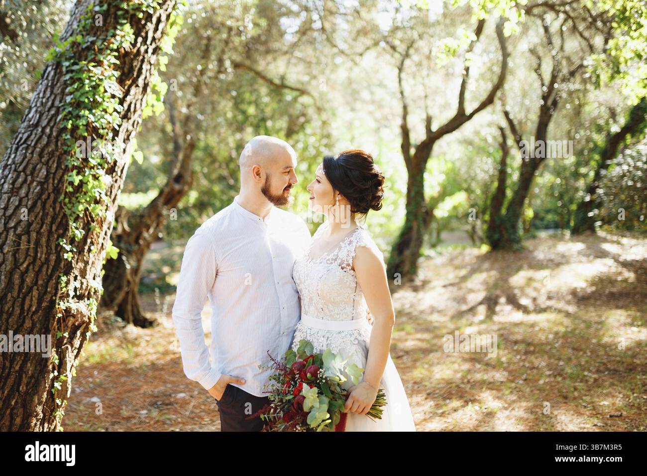 The bride and groom with a bouquet stand hugging among the trees in an ...