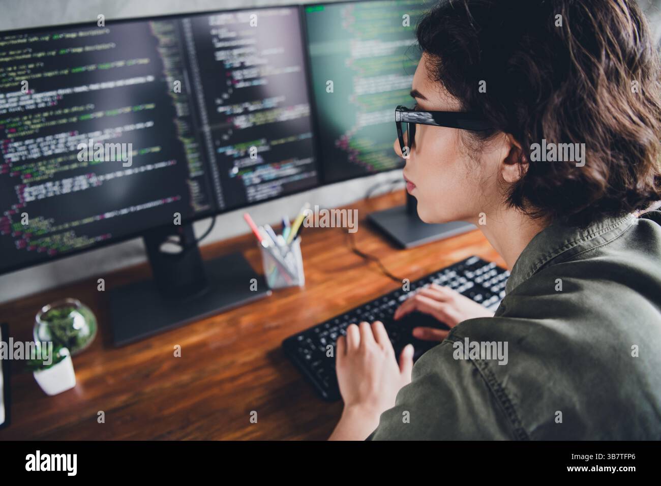 Young female programmer working on code at a modern workspace indoors using multiple screens, focused on software development Stock Photo