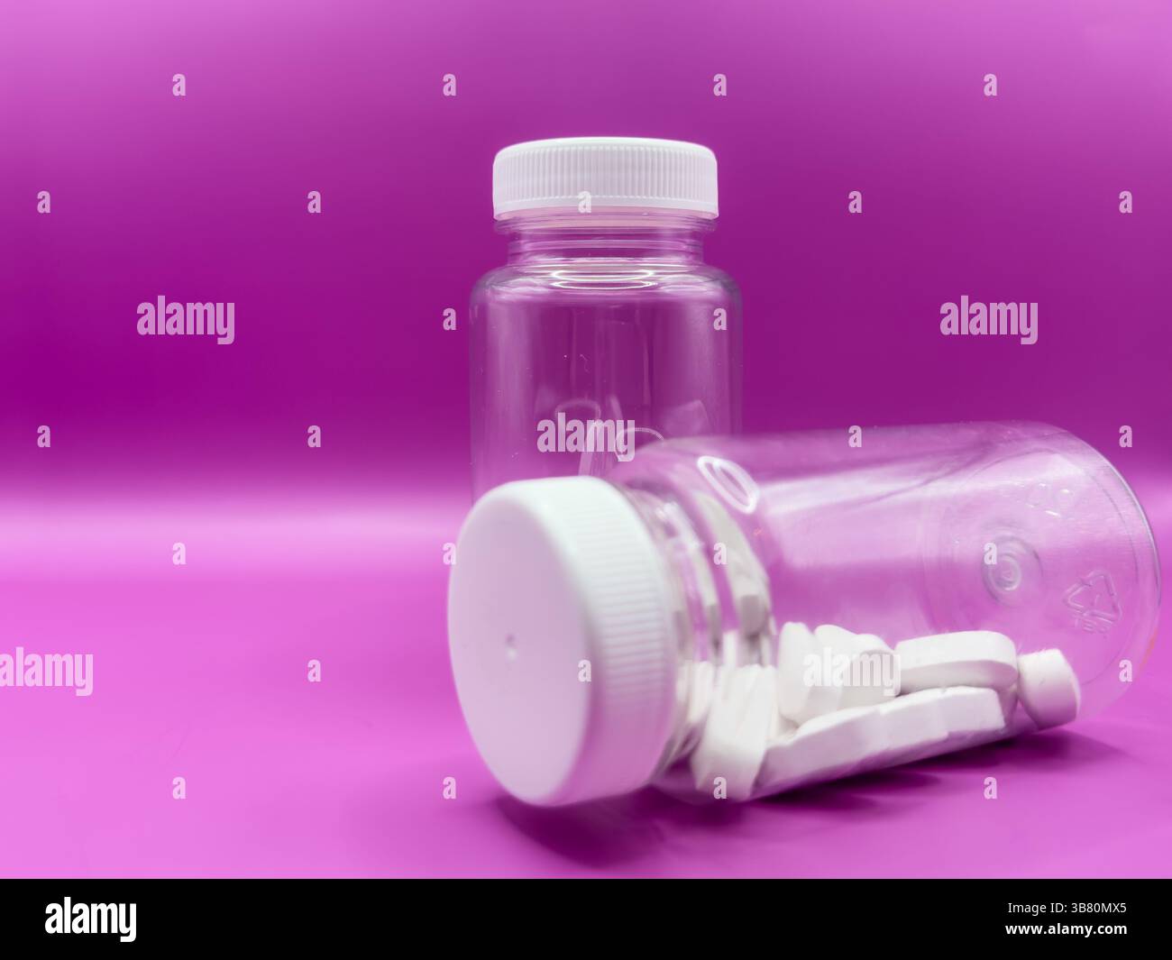 Two transparent jars with white pills, one jar is in the foreground, on a pink background, front view. Stock Photo