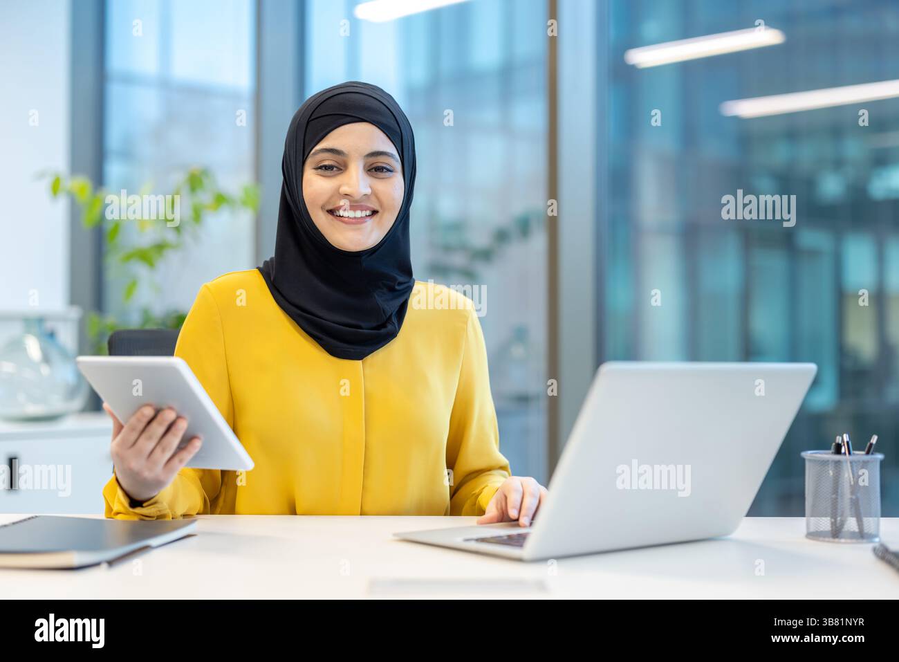 A smiling woman wearing a hijab works at her desk in a modern office ...