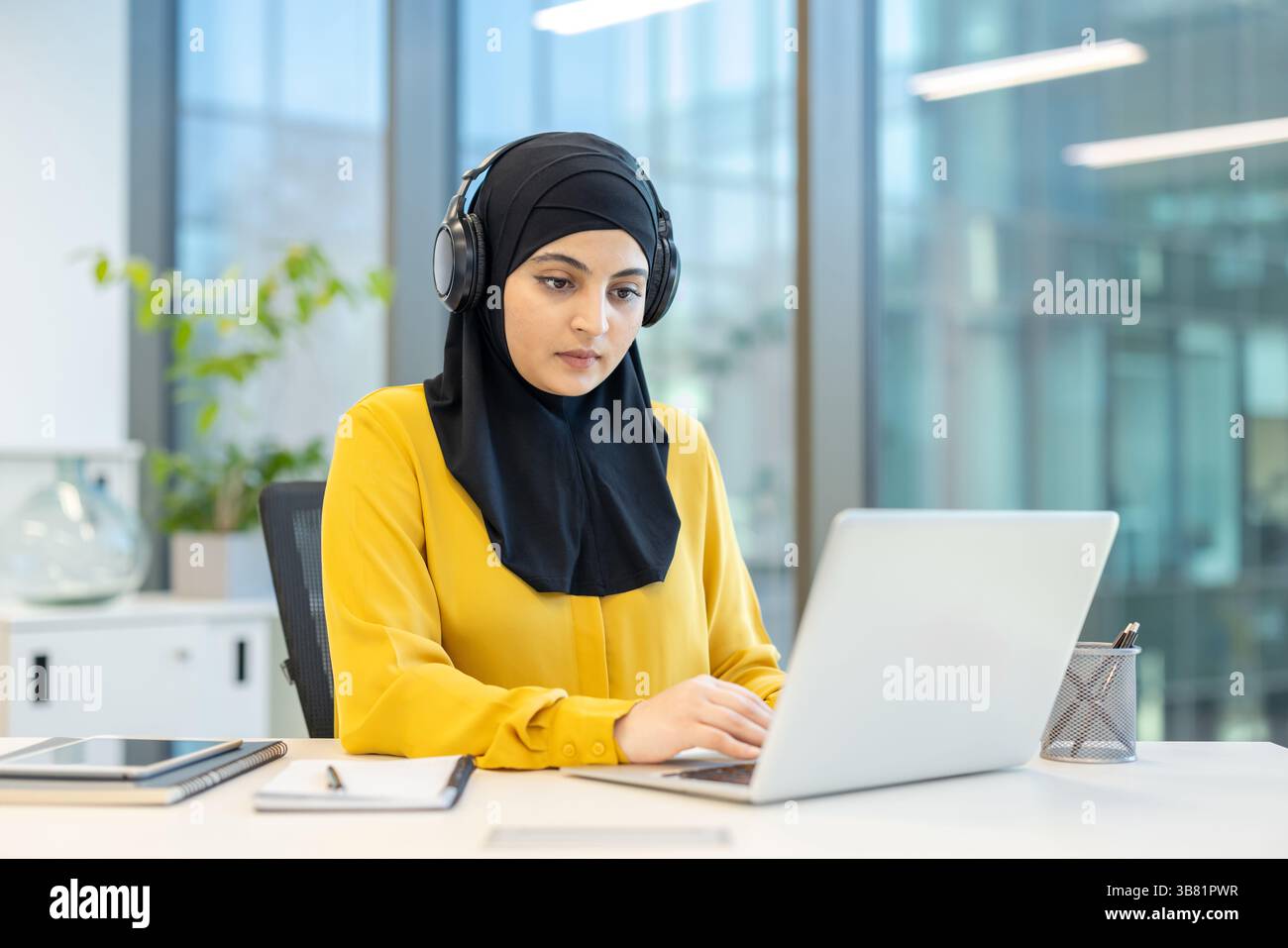 A woman wearing a hijab and headphones is working on her laptop in a ...