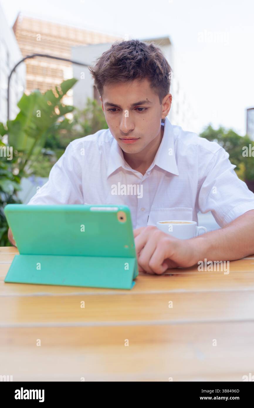 Focused young man using tablet at outdoor cafe table in vertical urban setting Stock Photo