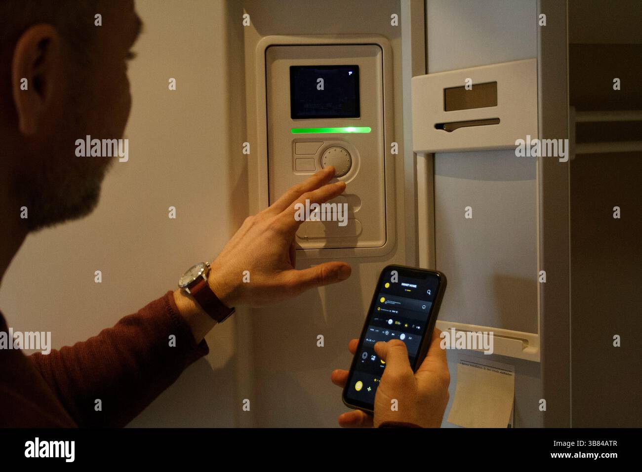 Man adjusting temperature through thermostat while holding smart phone Stock Photo