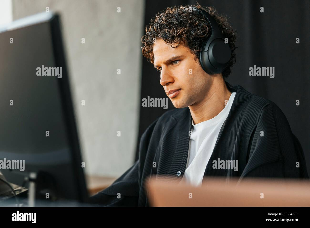 Curly haired young male computer programmer wearing wireless headphones using desktop PC in startup office Stock Photo