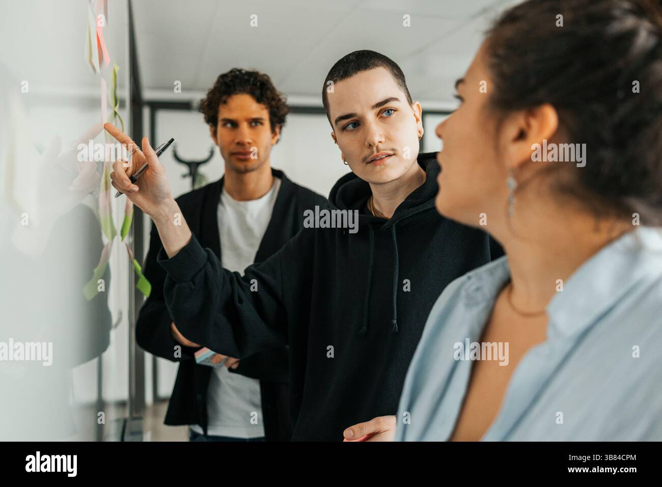 Non-binary business professional pointing on whiteboard while talking with colleague during discussion in office Stock Photo