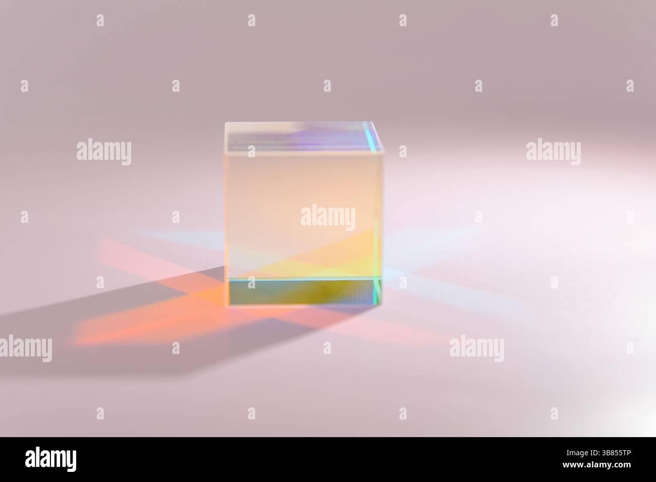 A clear prism refracts light, creating a vibrant display of gradient colors on a light pink surface, showcasing light properties Stock Photo