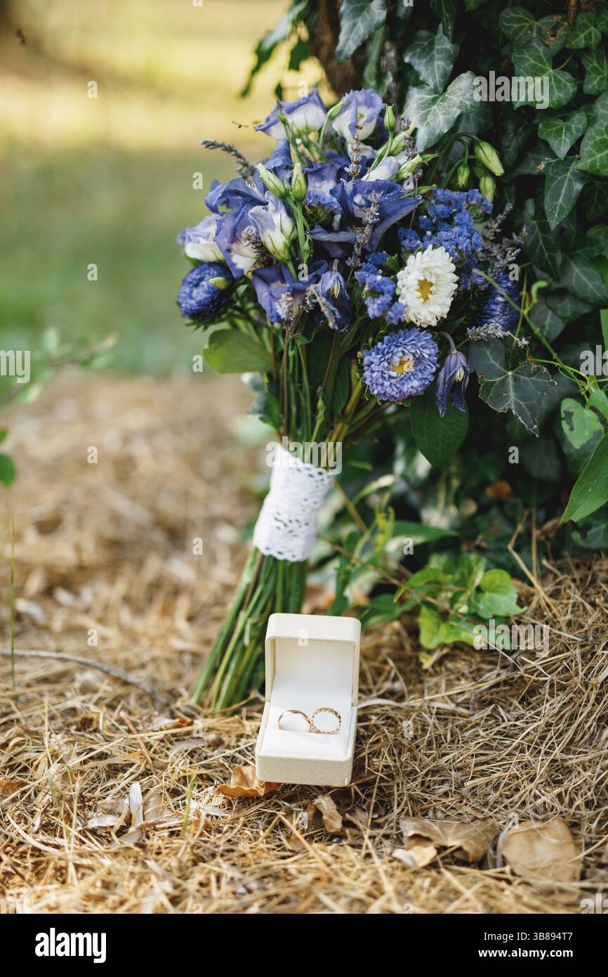 Wedding rings in a white box on dry grass with a bouquet of blue ...