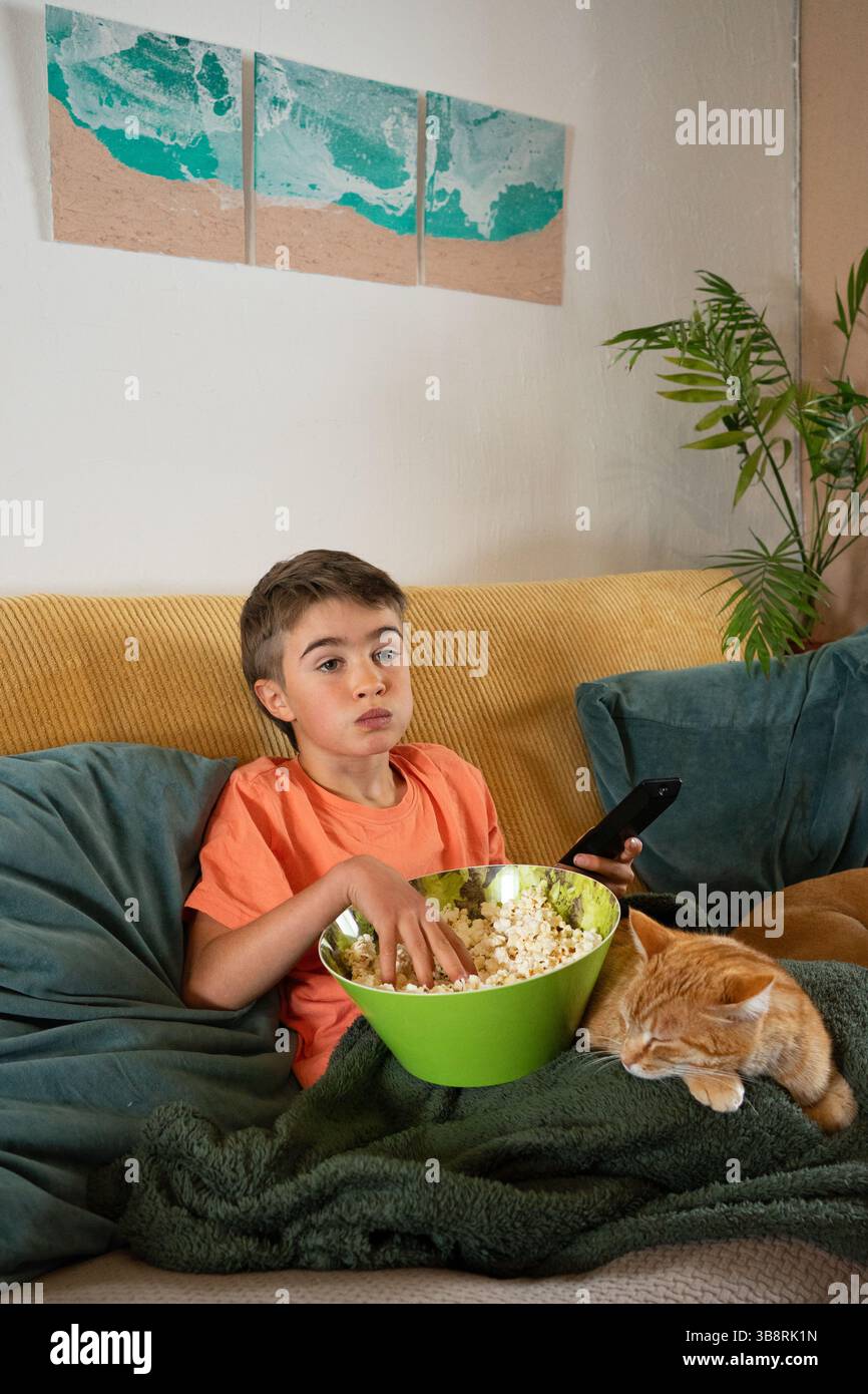 Boy eating popcorn watching tv on sofa with sleeping cat Stock Photo