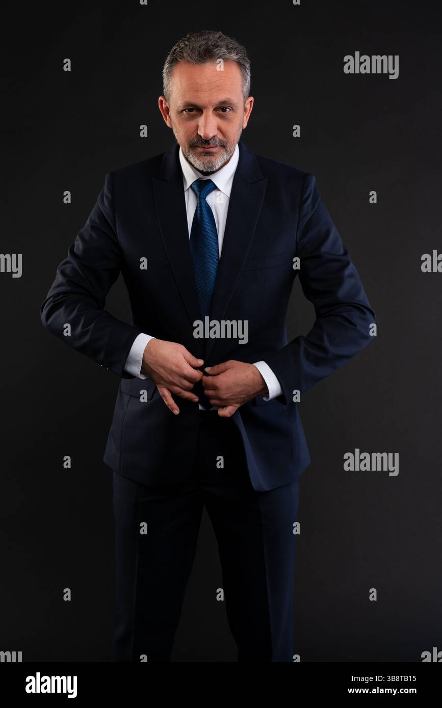 Mature businessman adjusting his formal outfit, buttoning his navy suit jacket while looking confidently at the camera. Stock Photo