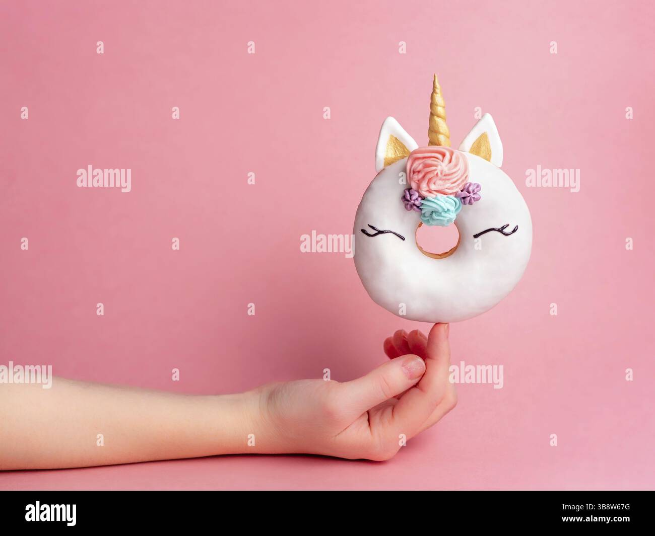 Unicorn donut on forefinger over pink background. Female hand hold trendy donut unicorn with white glaze on index finger. Copy space for text Stock Photo