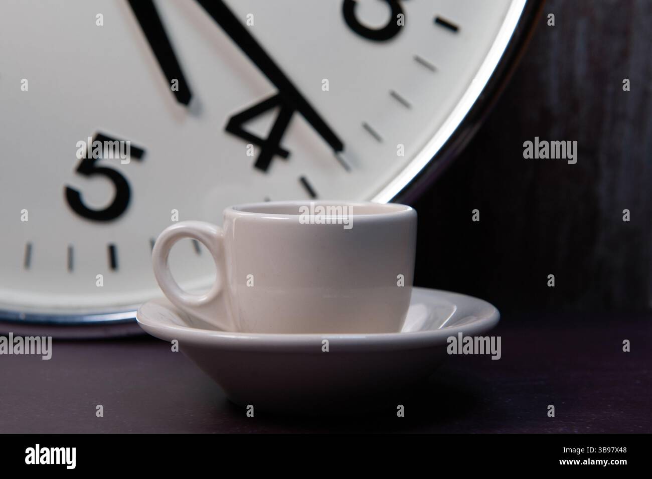 A white ceramic cappuccino cup and saucer in front of a large industrial clock. Stock Photo