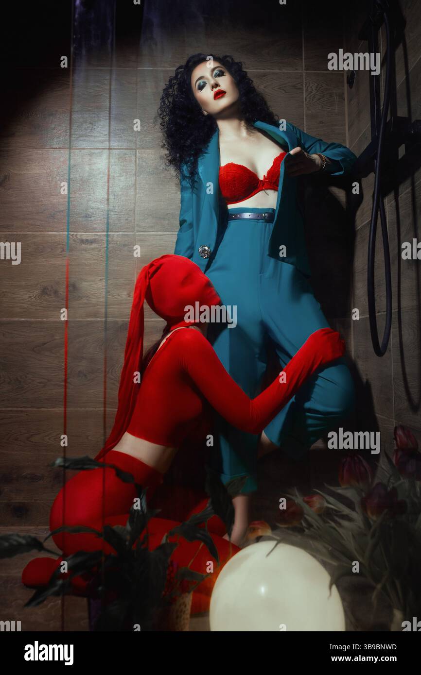 A woman in a teal outfit sits confidently as a red-costumed figure kneels beside her. A powerful, symbolic photo full of tension and meaning. Stock Photo