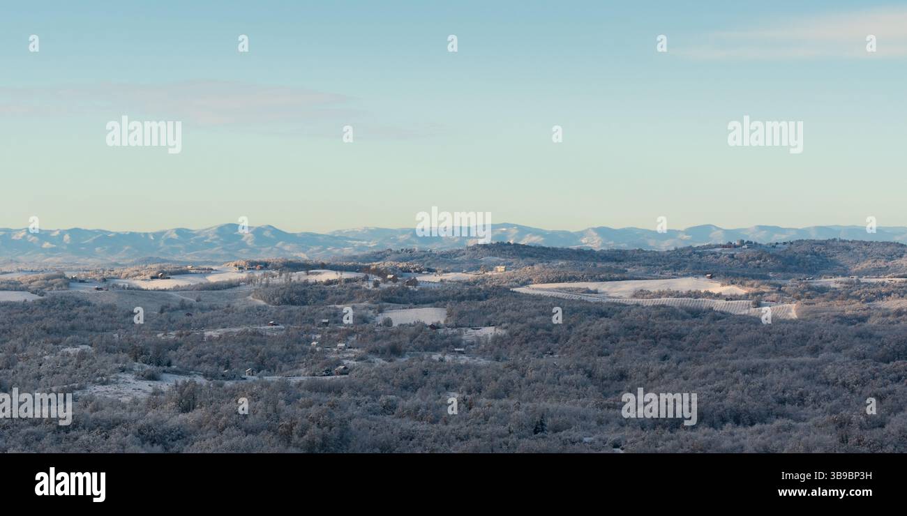 Hilly village covered with frost after cold winter night, rural landscape with houses, deciduous forest and fields, mountain range in distance Stock Photo
