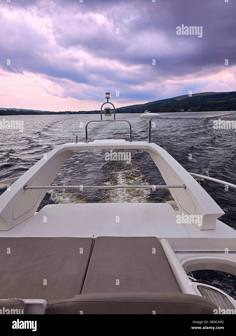 A sleek white boat glides across a rippling Loch Lomond, viewed from its stern. Moody purple-gray clouds hover above a distant mountain shoreline. - Smartphone Captured Stock Image