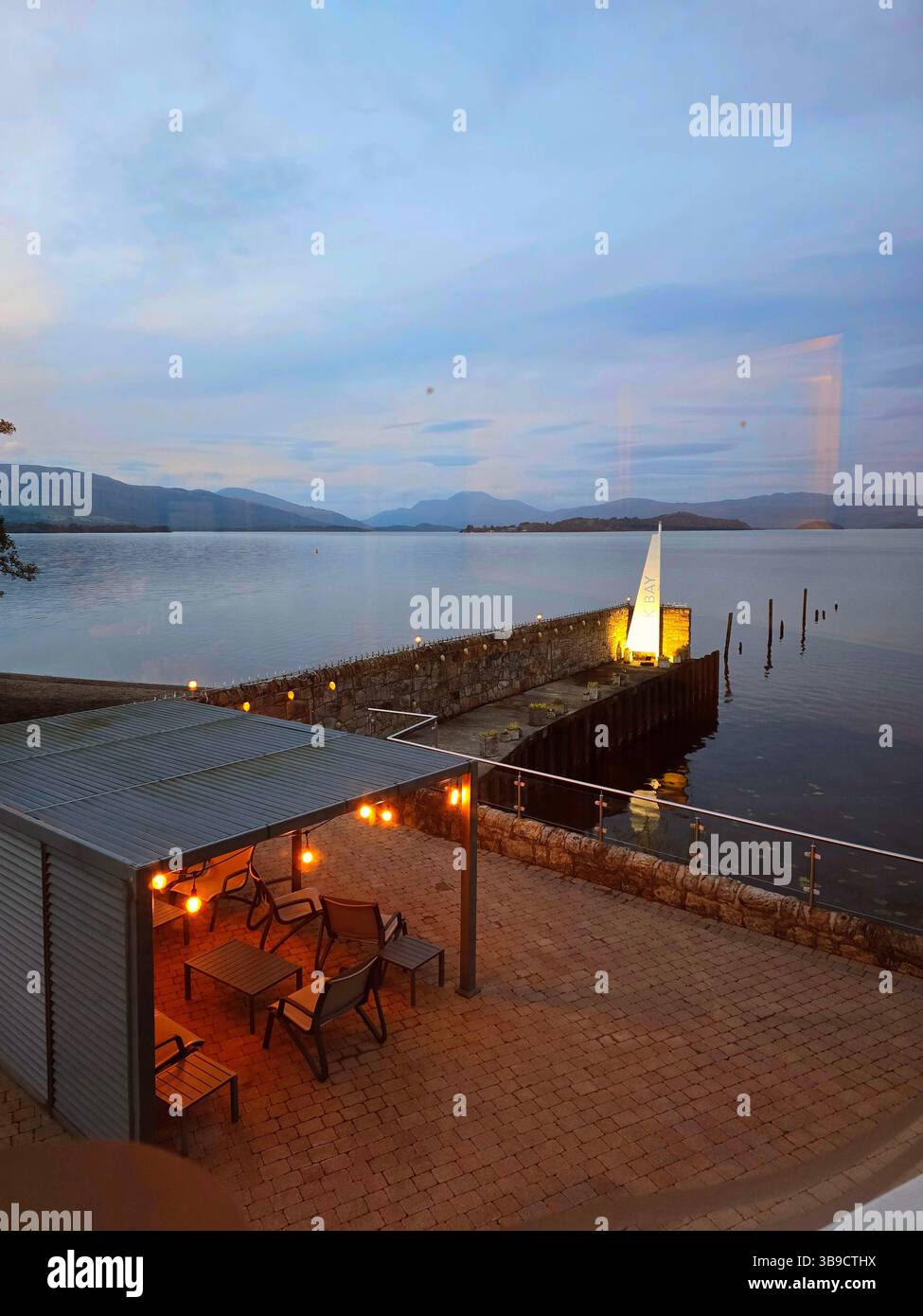 The Loch Lomond peaceful waterside retreat at dusk, with cozy lights and inviting chairs. An idyllic setting of the tranquil lake and mountains. - Smartphone Captured Stock Image