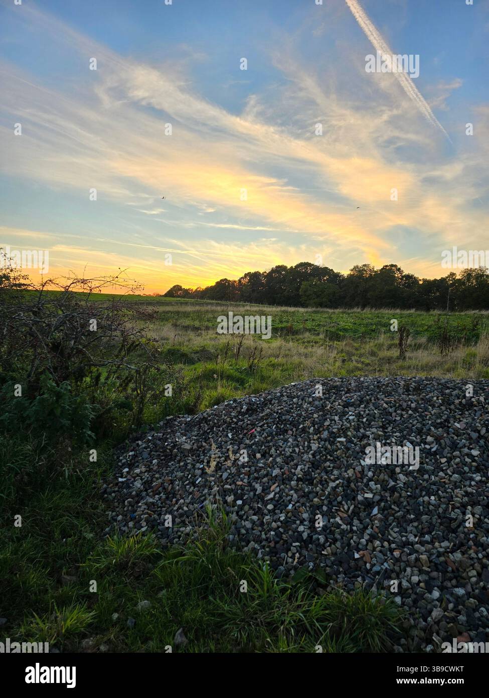 A serene countryside landscape at sunset, featuring gravel in the foreground, open grassy fields, and a vibrant sky painted with wispy clouds. - Smartphone Captured Stock Image