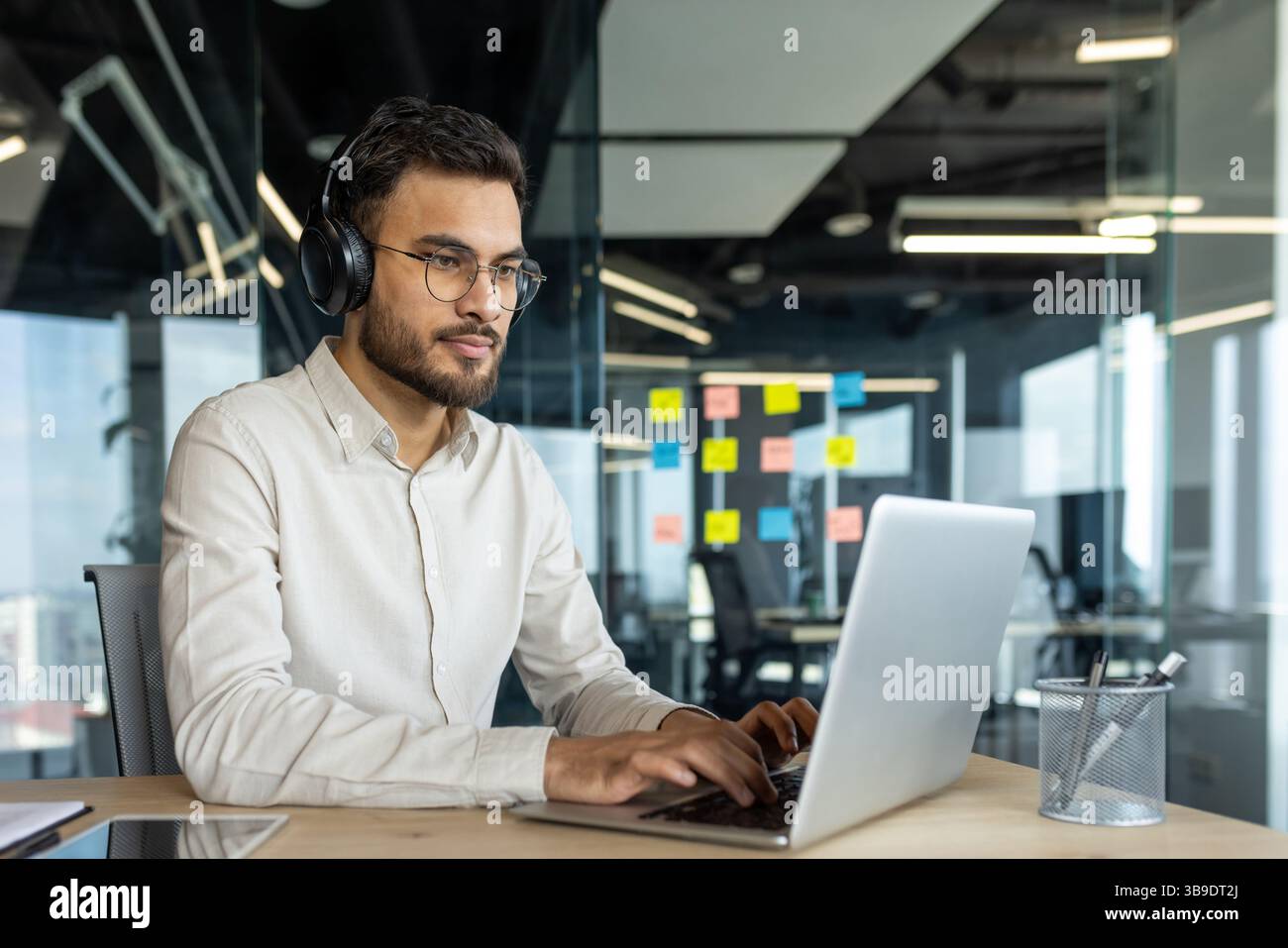 Serious thinking programmer coding new software. Man in headphones working with laptop inside office. Developer typing on keyboard. Stock Photo