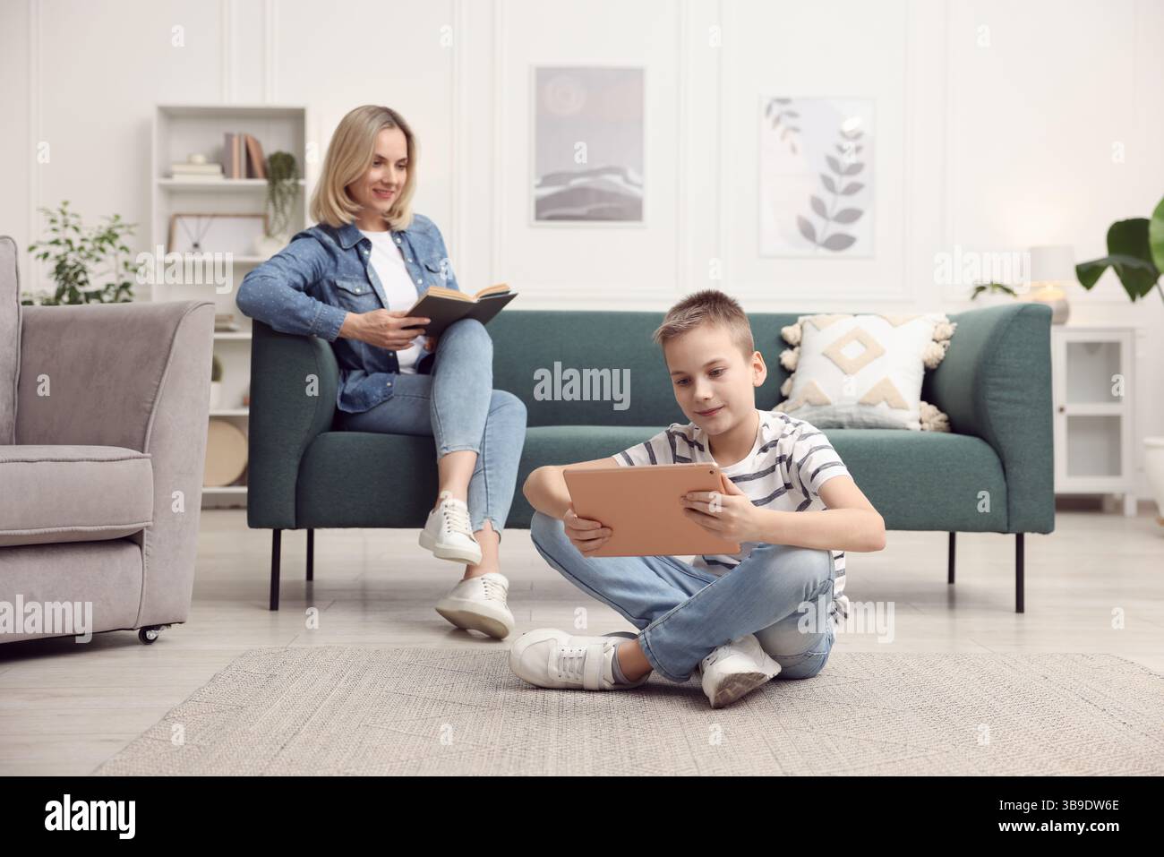Son using tablet while his mother reading book at home, selective focus Stock Photo