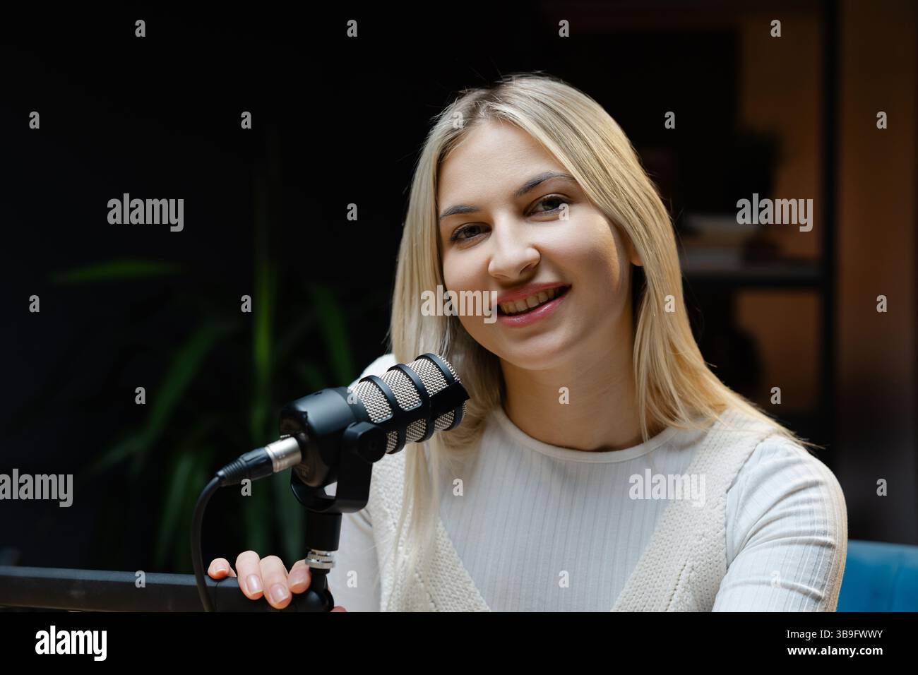 Young woman recording a podcast in her home studio, smiling and engaging with the microphone while sharing her thoughts and ideas Stock Photo