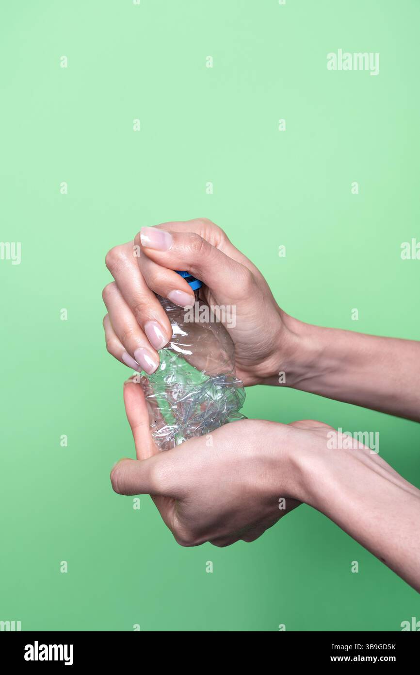 Two hands squeeze a clear plastic bottle, demonstrating recycling and waste reduction. The vibrant green background highlights the environmental focus Stock Photo