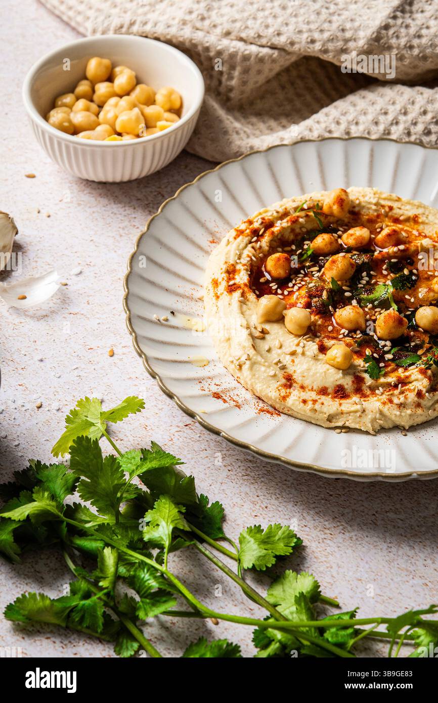 A plate of creamy hummus garnished with chickpeas, herbs, and spices sits on a textured background. A bowl of chickpeas and fresh herbs enhance the vi Stock Photo