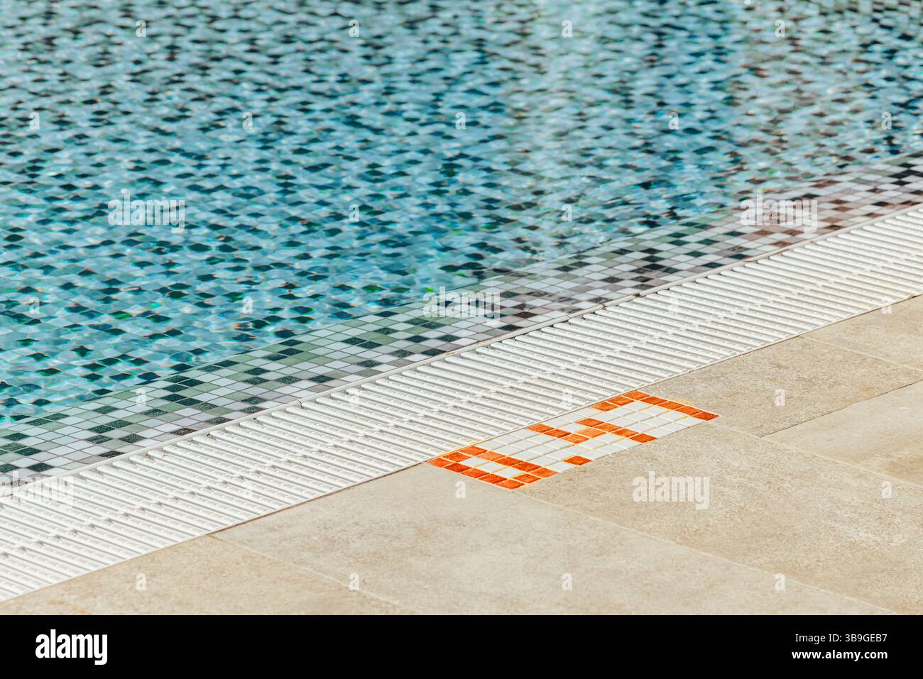 The image shows a swimming pool with clear blue water, bordered by a distinct checkered mosaic tile pattern. The edge features safety tiles and bright Stock Photo