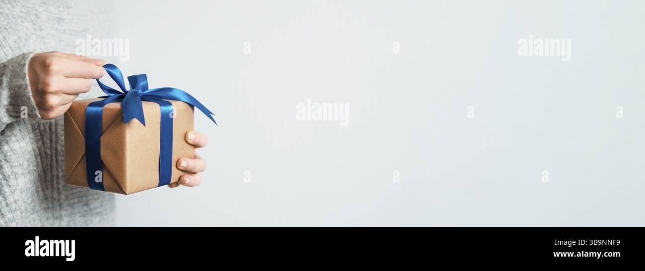 Female hands opening gift box on white background, copy space right.Unrecognizable caucasian girl in gray sweater holding gift box in craft wrapping p Stock Photo