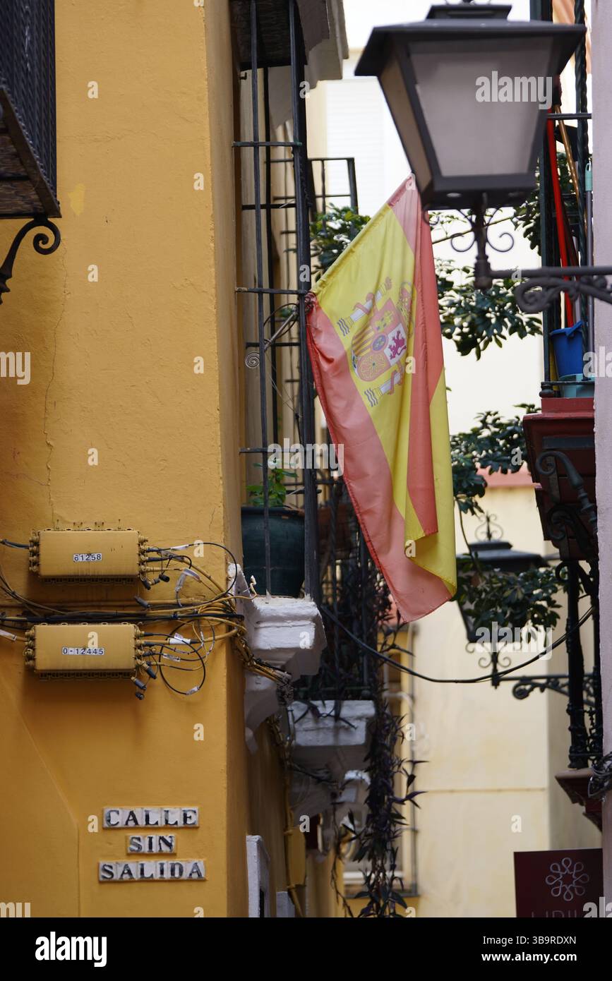 Spanish flag hanging in a narrow street in Seville, Spain, with a traditional lantern, ironwork details, and warm-coloured historic facades. Stock Photo