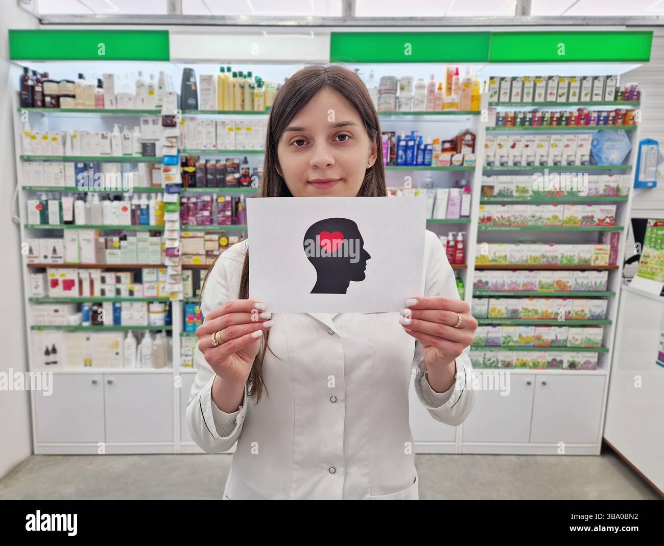 Female pharmacist holding a paper with a head silhouette featuring a heart inside, symbolizing mental health and wellness, over pharmacy shelves backd - Smartphone Captured Stock Image