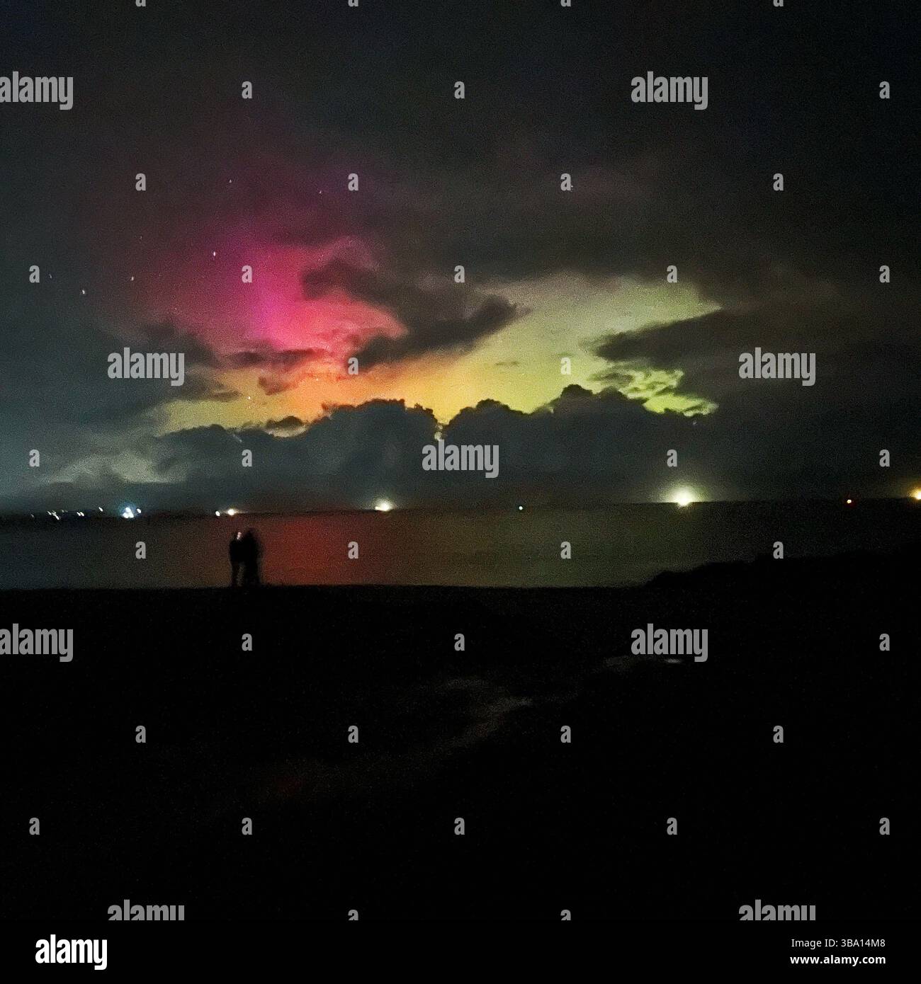 Silhouetted couple on Penmon Point beach under aurora borealis. Pink, yellow, orange and green lights above sea. Taken November 5 2023. - Smartphone Captured Stock Image