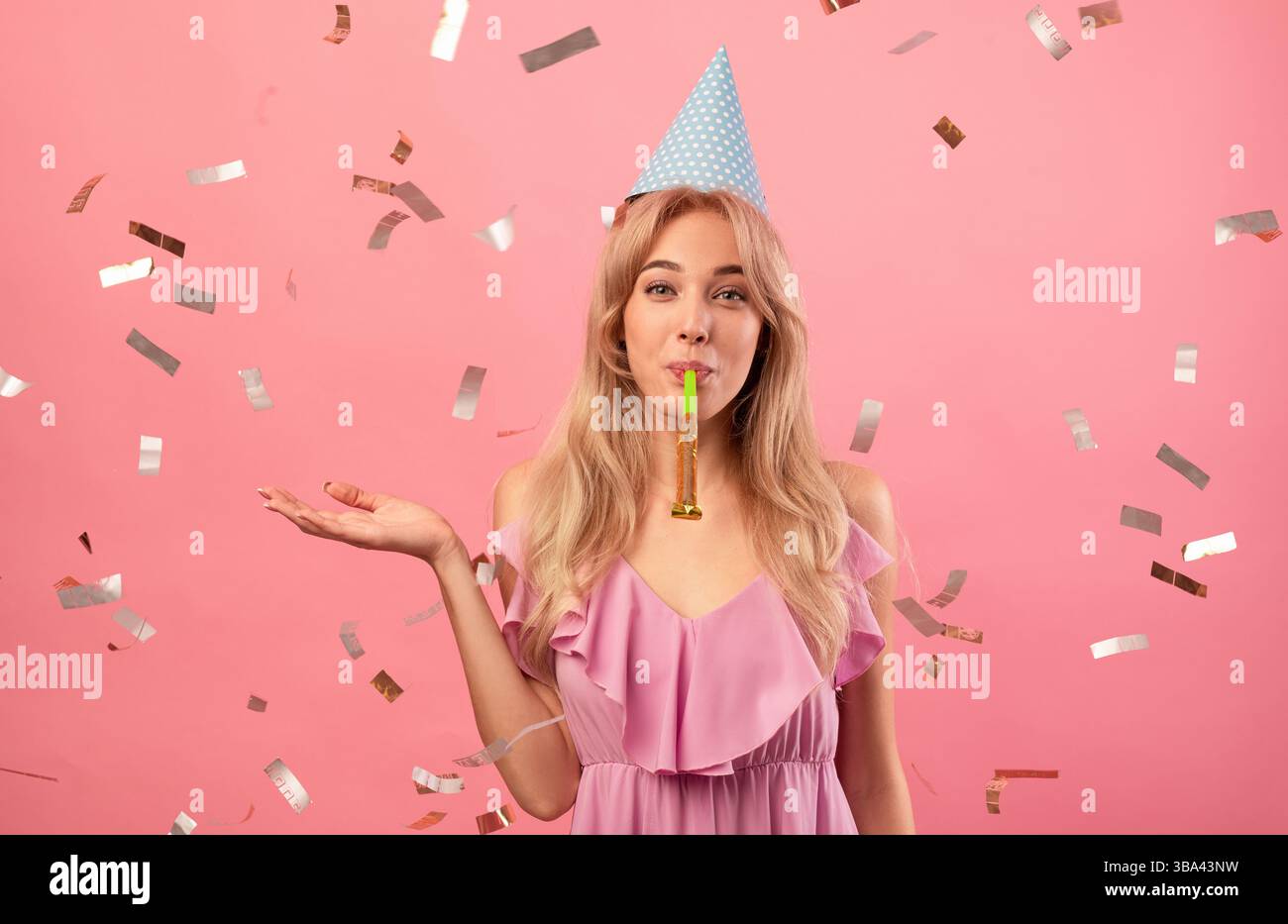 Gorgeous young woman in birthday cap blowing party horn on pink studio background with falling confetti Stock Photo