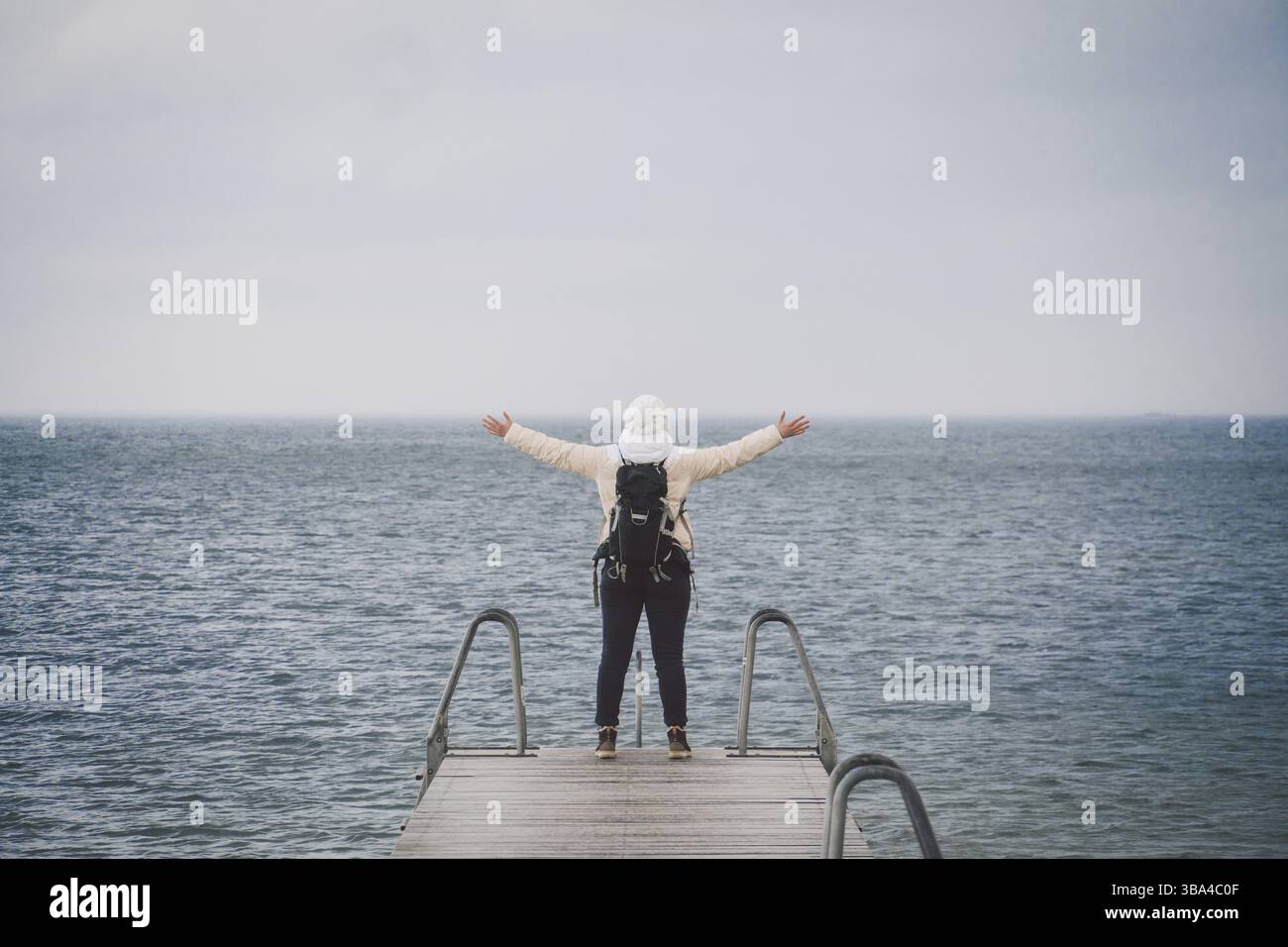 Concept of freedom relaxation. woman relaxing on pier. Backpacker hiker woman with backpack on sea wooden pier stands arms sideways looking to horizon Stock Photo