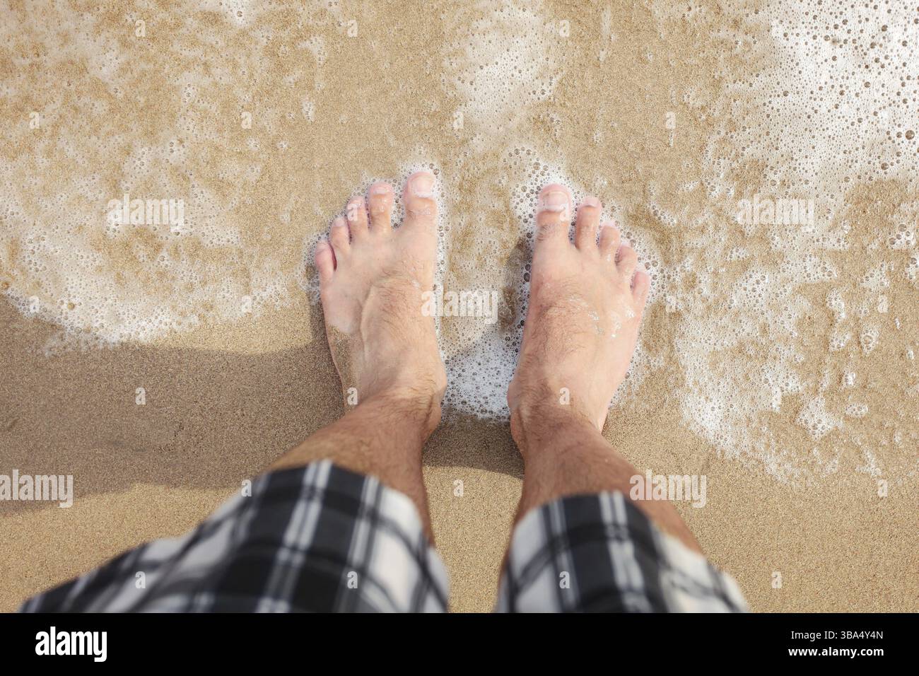 Looking down to man in shorts feet standing at wet beach sand, sea washing them, sun shining on, Dipkarpaz, Cyprus, Europe Stock Photo