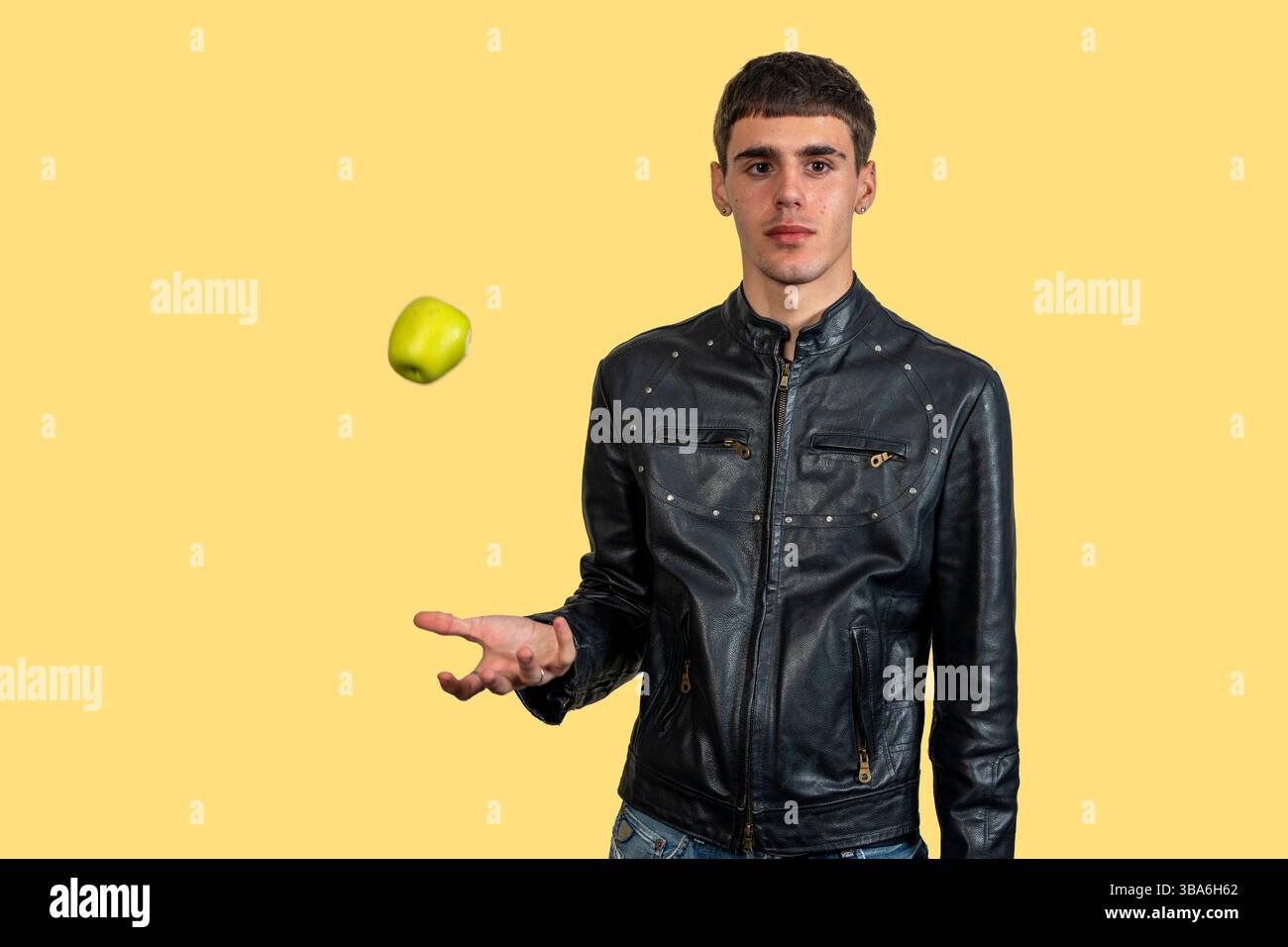 Young man wearing black leather jacket juggling green apple on yellow background Stock Photo