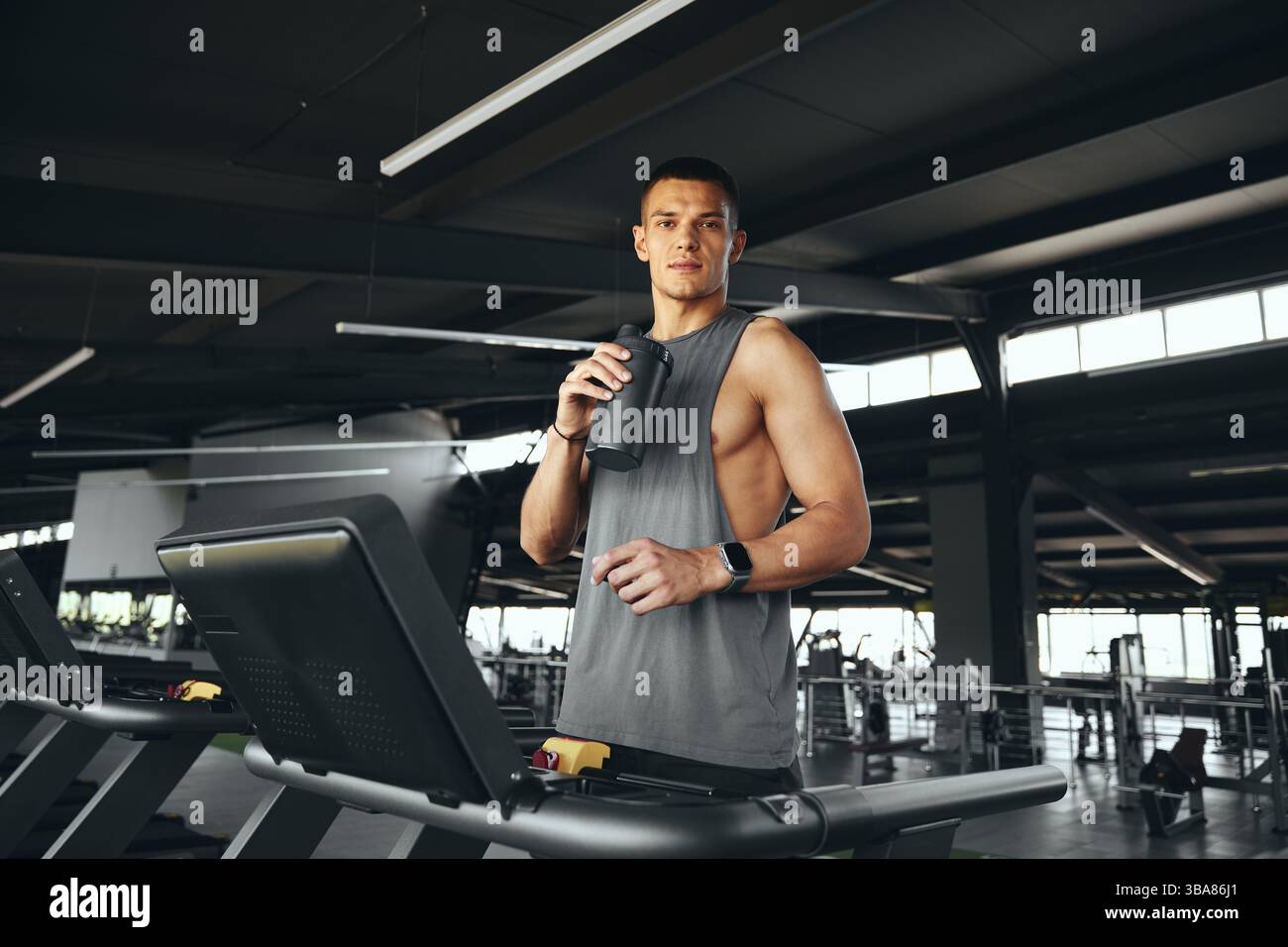 Active young guy in grey tshirt drinking water from bottle, standing on treadmill. Stock Photo
