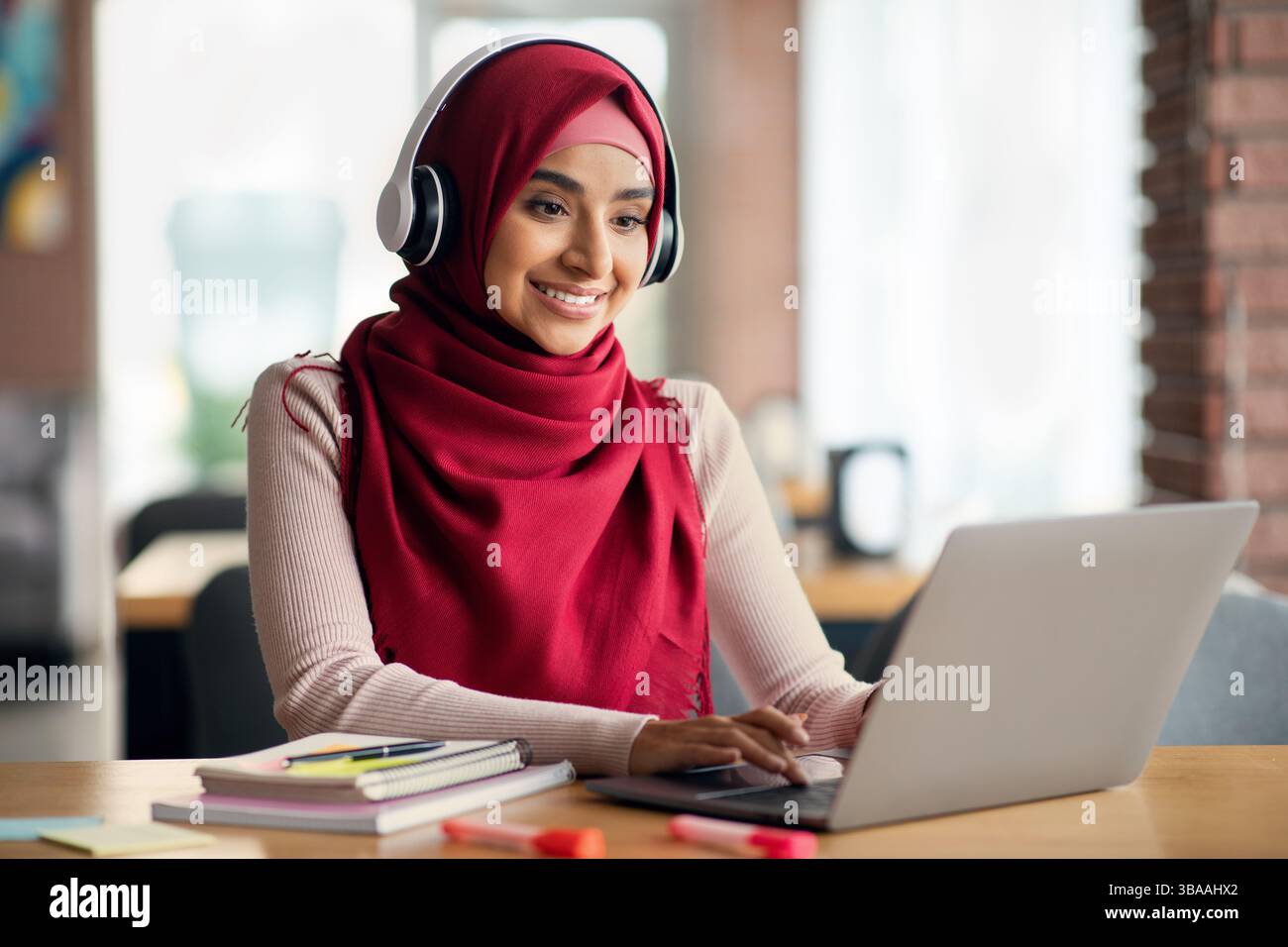 Joyful young muslim woman in hijab sitting at table in front of laptop ...