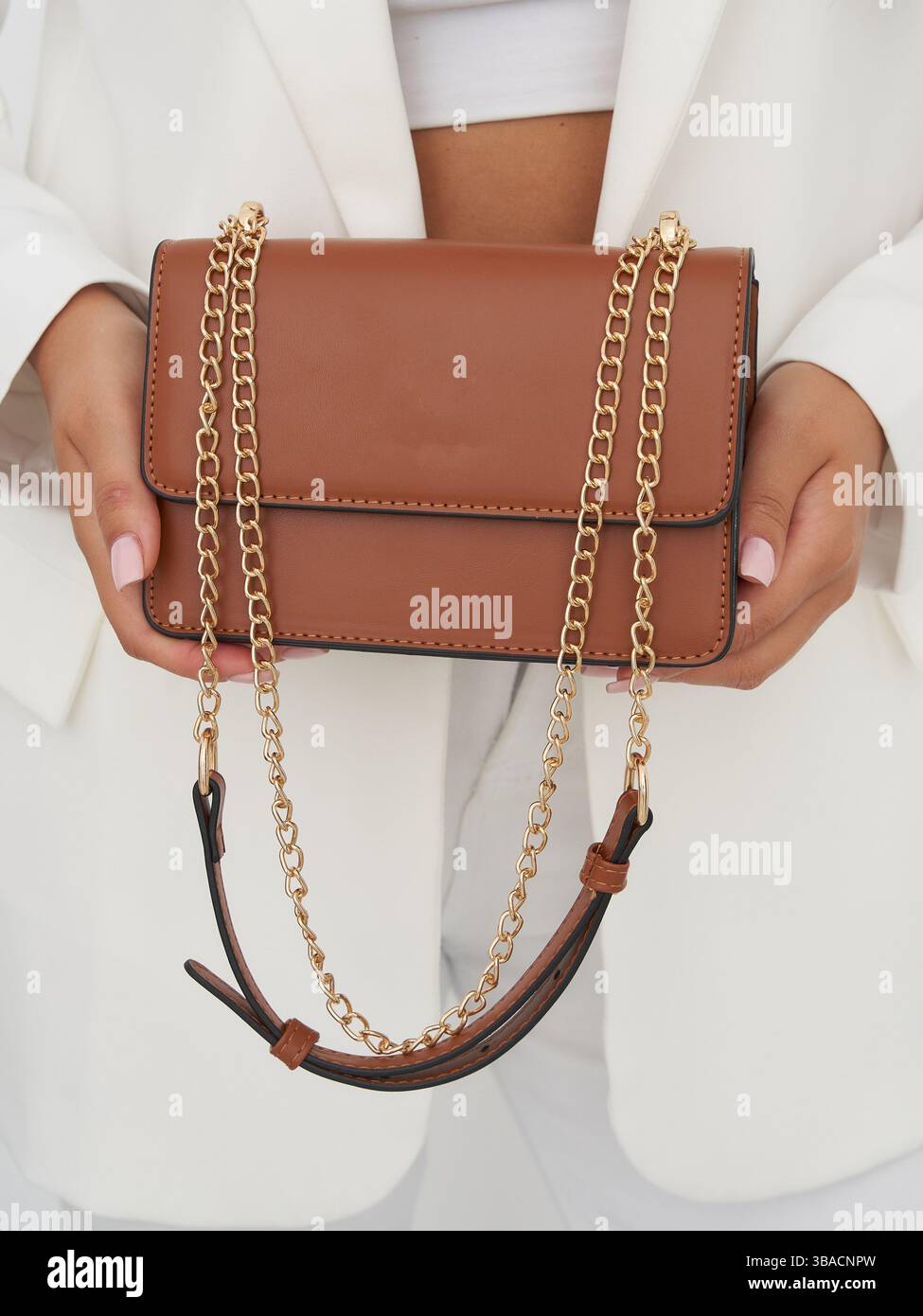 Elegant brown handbag with gold chain strap held by individual wearing a white suit and a white top. The handbag features a sleek design with fine Stock Photo