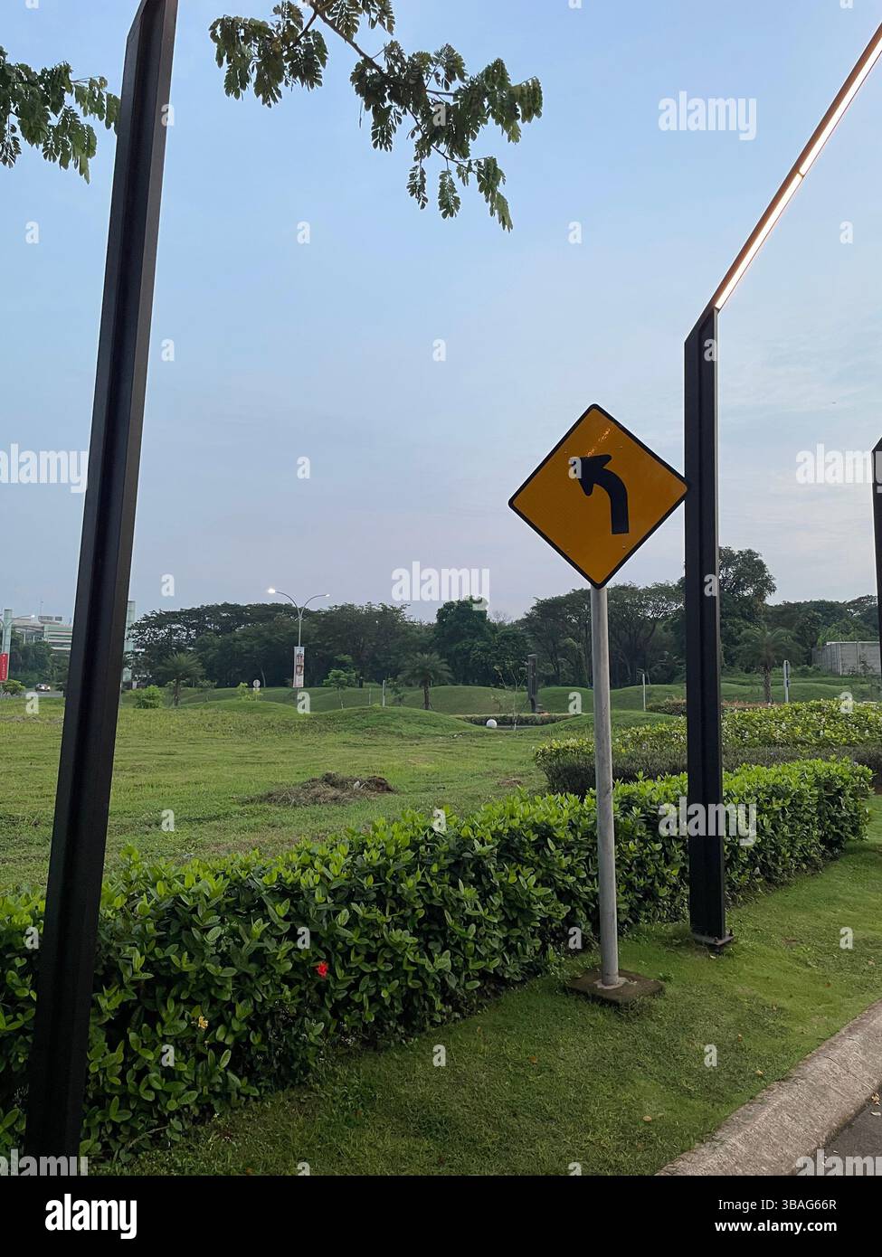 A yellow left turn traffic sign positioned beside a manicured green roadside with metal poles and blue sky in the background. - Smartphone Captured Stock Image