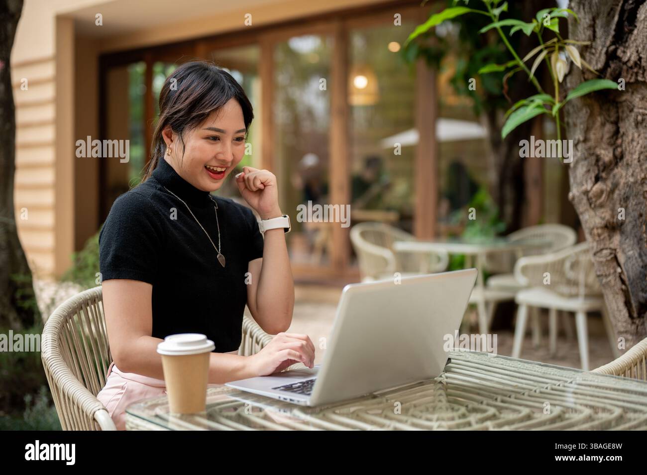 A cheerful, excited Asian woman sits at an outdoor table of a cafe ...