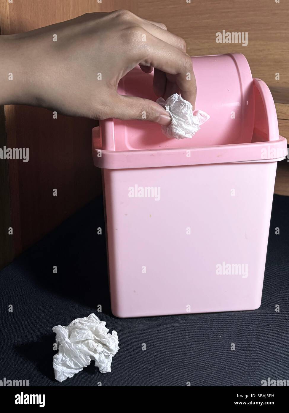 A crumpled tissue and paper are being tossed to a small desktop trash bin with flip-top lid. - Smartphone Captured Stock Image