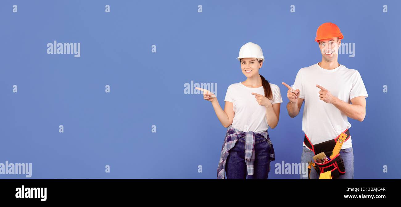 Smiling handyworkers couple wearing casual clothes and protective hard hats pointing aside Stock Photo