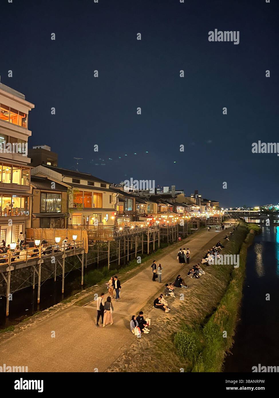 Evening view of people relaxing and dining along the Kamo River near Pontocho in Kyoto, Japan. Traditional buildings illuminated at night. - Smartphone Captured Stock Image