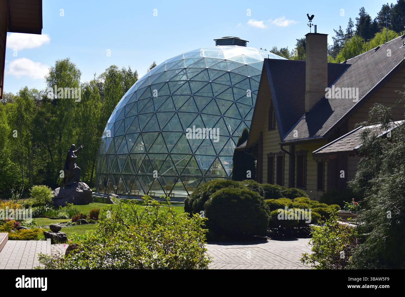 Glass geodesic dome surrounded by nature, next to a traditional wooden house. A statue stands in the garden, blending modern architecture Stock Photo