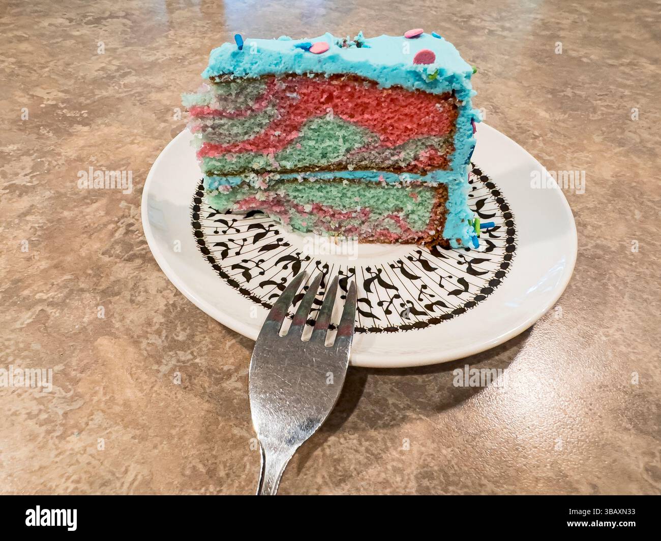 Multi-colored birthday cake slice, on a plate, with a well-used fork.  The counter surface is probably Formica. - Smartphone Captured Stock Image