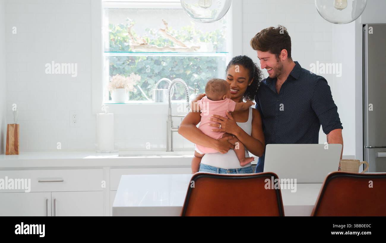 Family With Baby Daughter In Kitchen Using Laptop On Counter Stock Photo