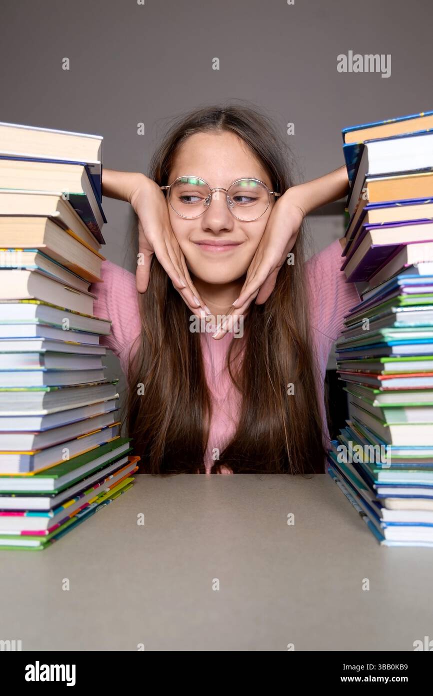 Thoughtful student sitting between stacks of books. A young student wearing glasses sits with her chin resting on her hands between two tall stacks of Stock Photo