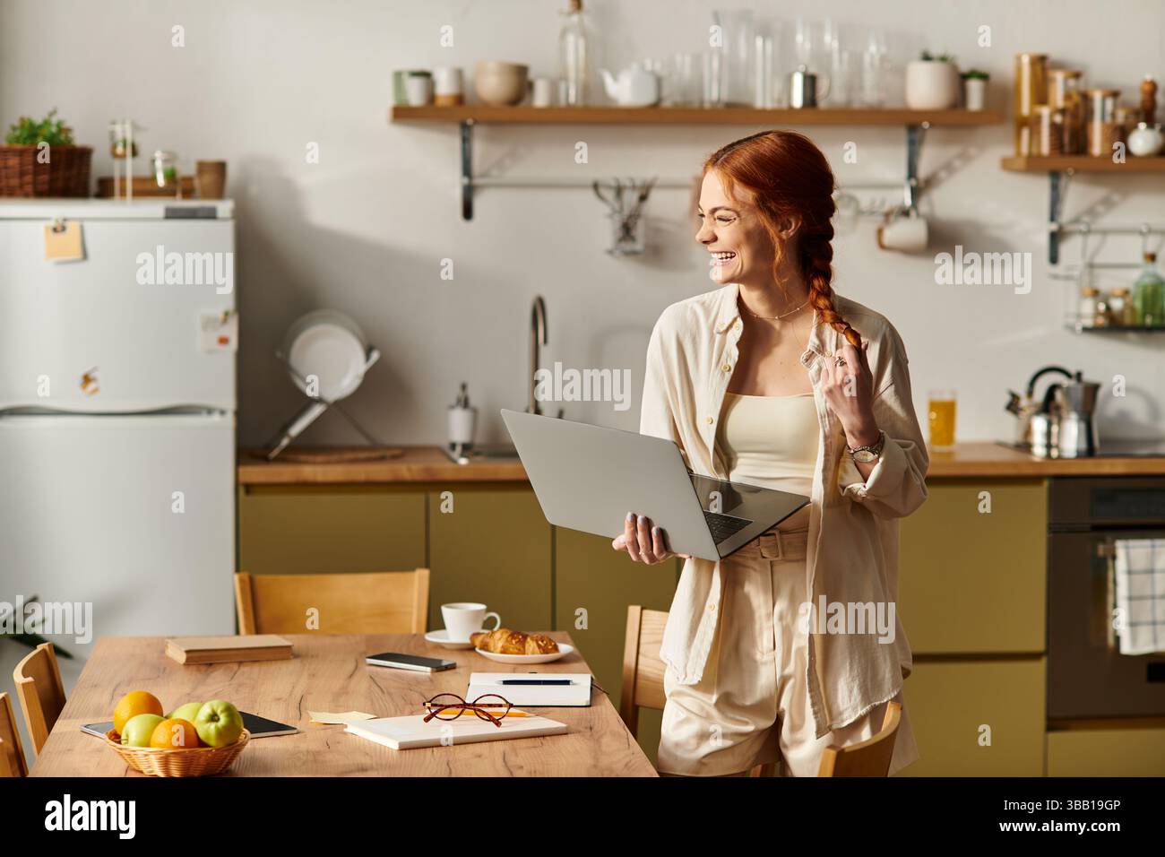 Brightly lit kitchen showcases a cheerful red haired woman engaged in creative work while smiling. Stock Photo
