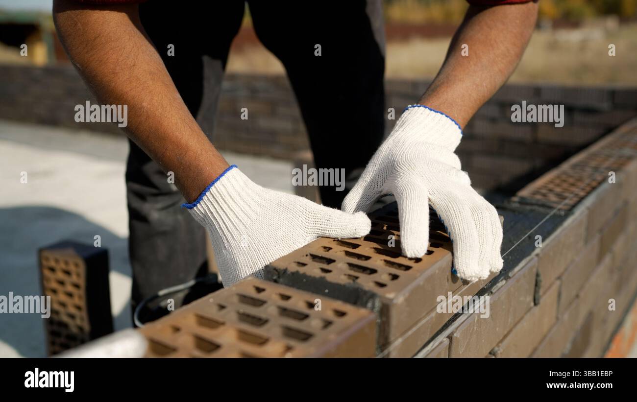 Construction worker carefully laying bricks on fresh cement, using precise hammer and level techniques while building sturdy wall section with profess Stock Photo